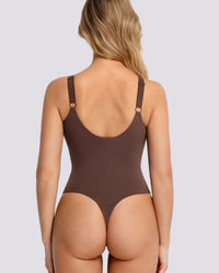 Rear view of a woman wearing a brown one-piece bodysuit with thin shoulder straps and small gold-tone ring details, against a light gray studio background.