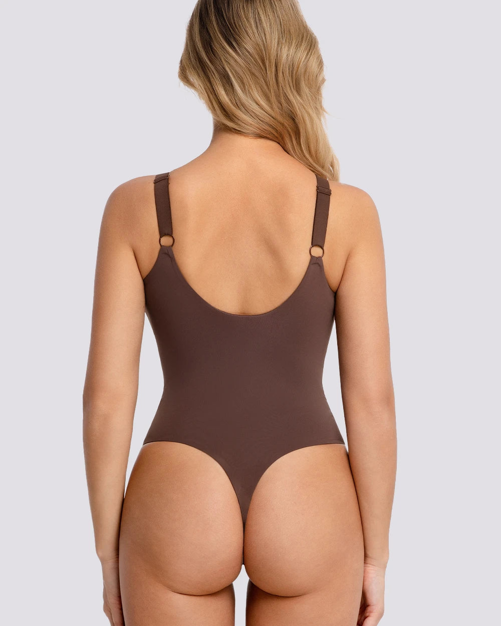 Rear view of a woman wearing a brown one-piece bodysuit with thin shoulder straps and small gold-tone ring details, against a light gray studio background.
