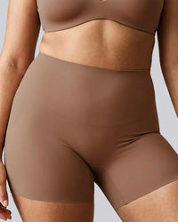 Close-up of a model wearing high-waisted shapewear shorts in a solid taupe-brown color, paired with a matching bralette, against a neutral gray background.