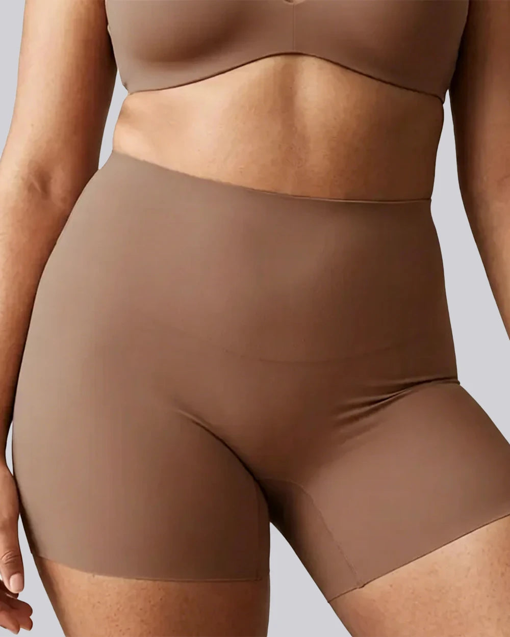 Close-up of a model wearing high-waisted shapewear shorts in a solid taupe-brown color, paired with a matching bralette, against a neutral gray background.