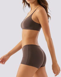 A woman wearing a matching two-piece activewear set in a dark taupe brown color, featuring a sports bra and high-waisted shorts, shown from a side profile against a light gray background.