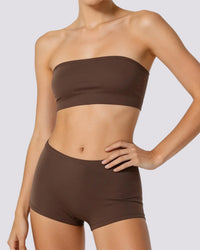 A woman wearing a matching dark brown strapless bandeau top and high-waisted shorts set, shown from the neck to the upper thighs against a light gray studio background.