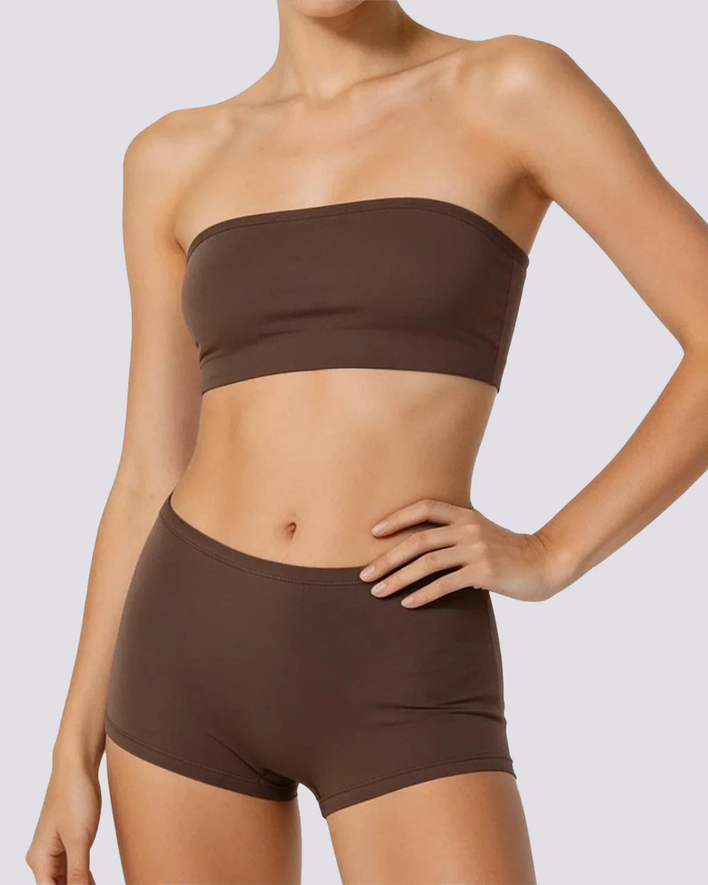 A woman wearing a matching dark brown strapless bandeau top and high-waisted shorts set, shown from the neck to the upper thighs against a light gray studio background.