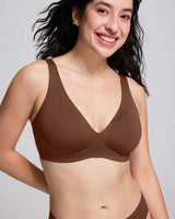 A smiling woman with dark hair wearing a solid brown bralette against a light gray studio background.