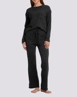 A woman modeling a dark charcoal ribbed knit lounge set consisting of a long-sleeve top and matching wide-leg pants, shown against a light gray studio background.