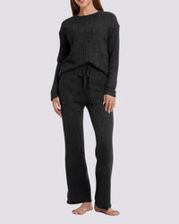 A woman modeling a dark charcoal ribbed knit lounge set consisting of a long-sleeve top and matching wide-leg pants, shown against a light gray studio background.
