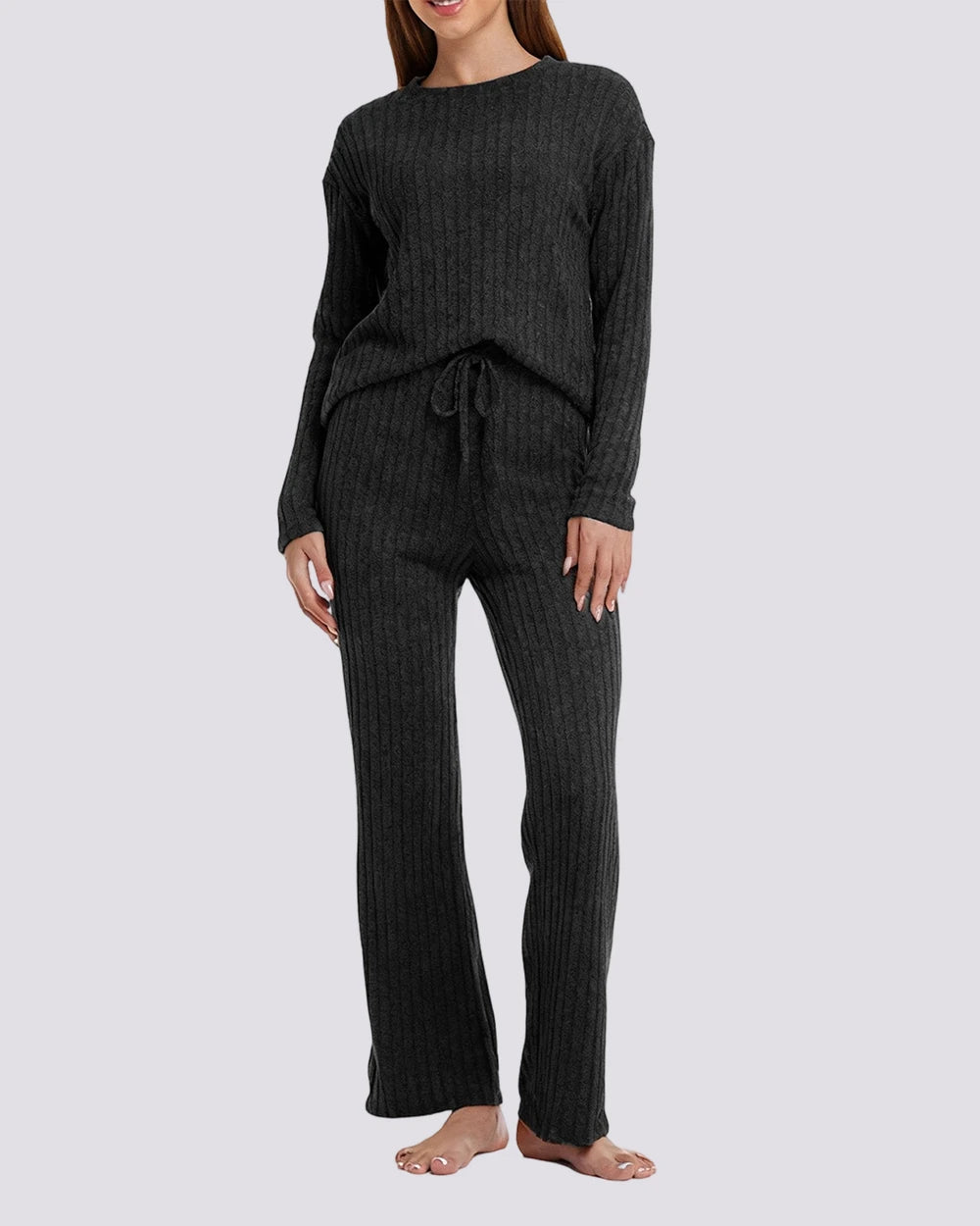 A woman modeling a dark charcoal ribbed knit lounge set consisting of a long-sleeve top and matching wide-leg pants, shown against a light gray studio background.