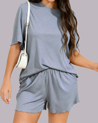 A woman wearing a matching two-piece lounge set consisting of a short-sleeve crewneck t-shirt and high-waisted shorts with scalloped hems, in a dusty slate blue color. She is carrying a small white shoulder bag against a solid gray studio background.