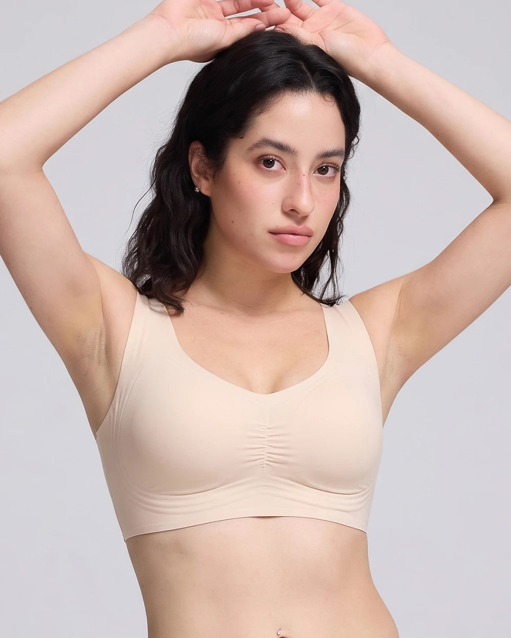 A woman modeling a beige wireless bra with a gathered, ruched center front, set against a solid light gray background.