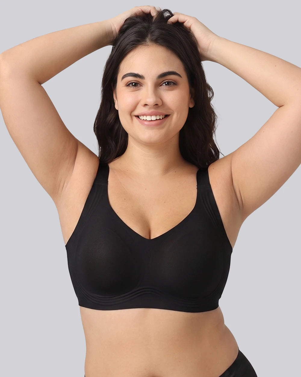 A woman with dark hair smiling and posing with her hands in her hair, wearing a black V-neck bralette against a solid gray background.