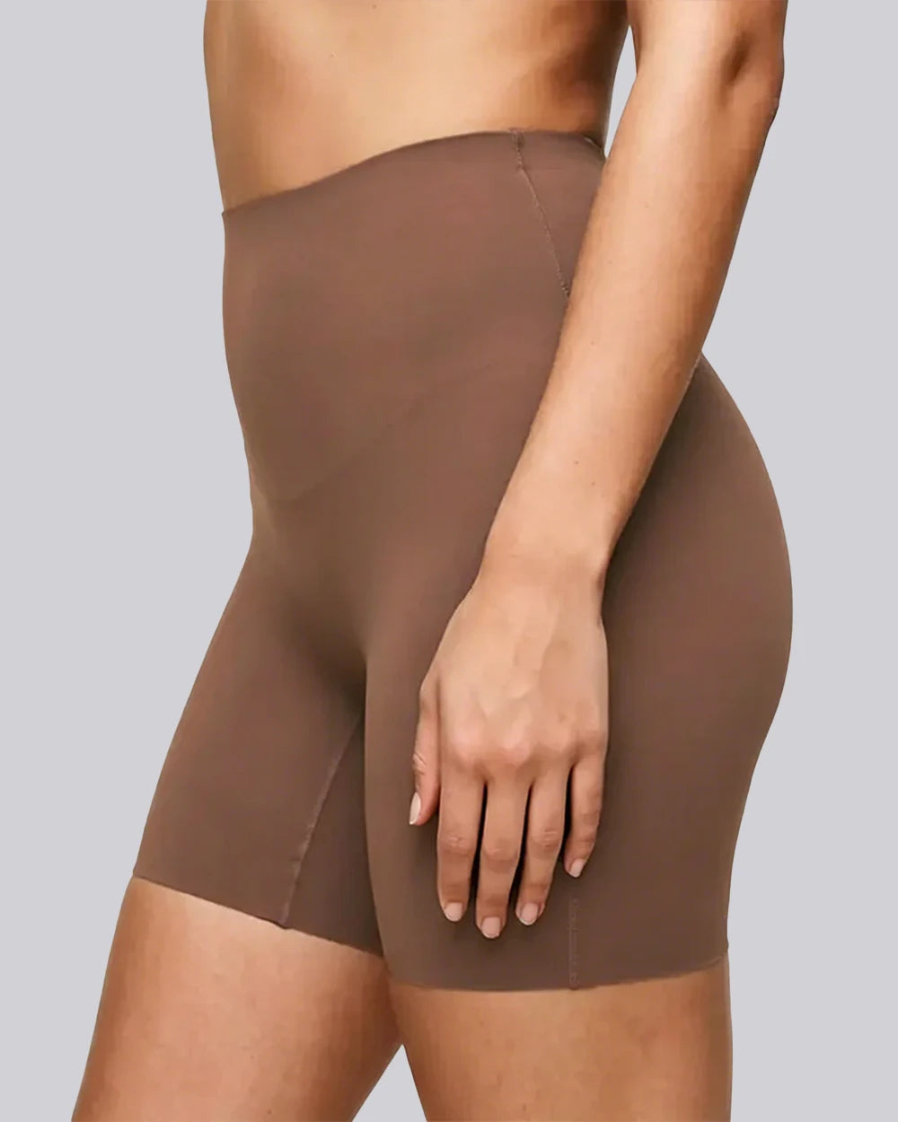 A close-up of a model wearing high-waisted, seamless brown shapewear shorts against a solid gray background.
