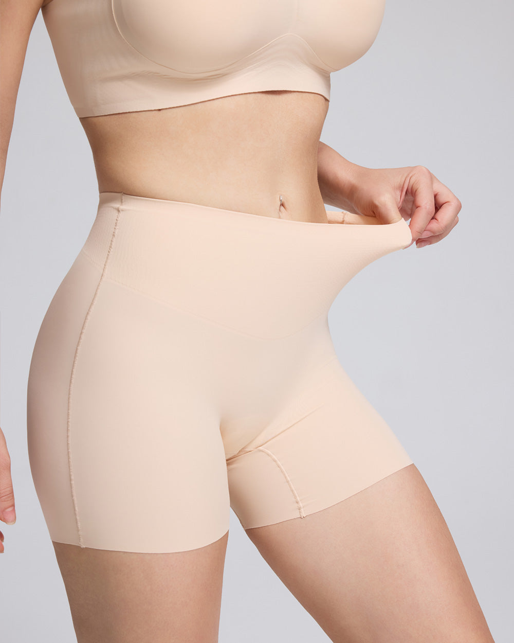 Close-up of a model wearing high-waisted shapewear shorts in a light nude beige color, pulling the waistband slightly outward to demonstrate stretch, against a solid light gray background.