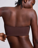 Close-up back view of a woman wearing a wide, strapless bandeau-style top in a muted mauve-brown color, with one hand pulling slightly at the band.
