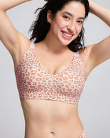 A woman wearing a pink and beige leopard-print bralette, posing with her arms raised against a light gray background.