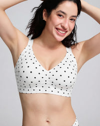A woman wearing a white V-neck bralette with black polka dots, posing with her arms raised against a light gray studio background.