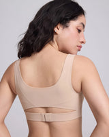 A woman shown from the back wearing a beige seamless bra with a wide underband and a small cutout detail, set against a plain light gray studio background.