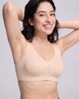 A smiling woman with dark hair wearing a beige wireless sports bra, posing with her arms raised against a light gray studio background.