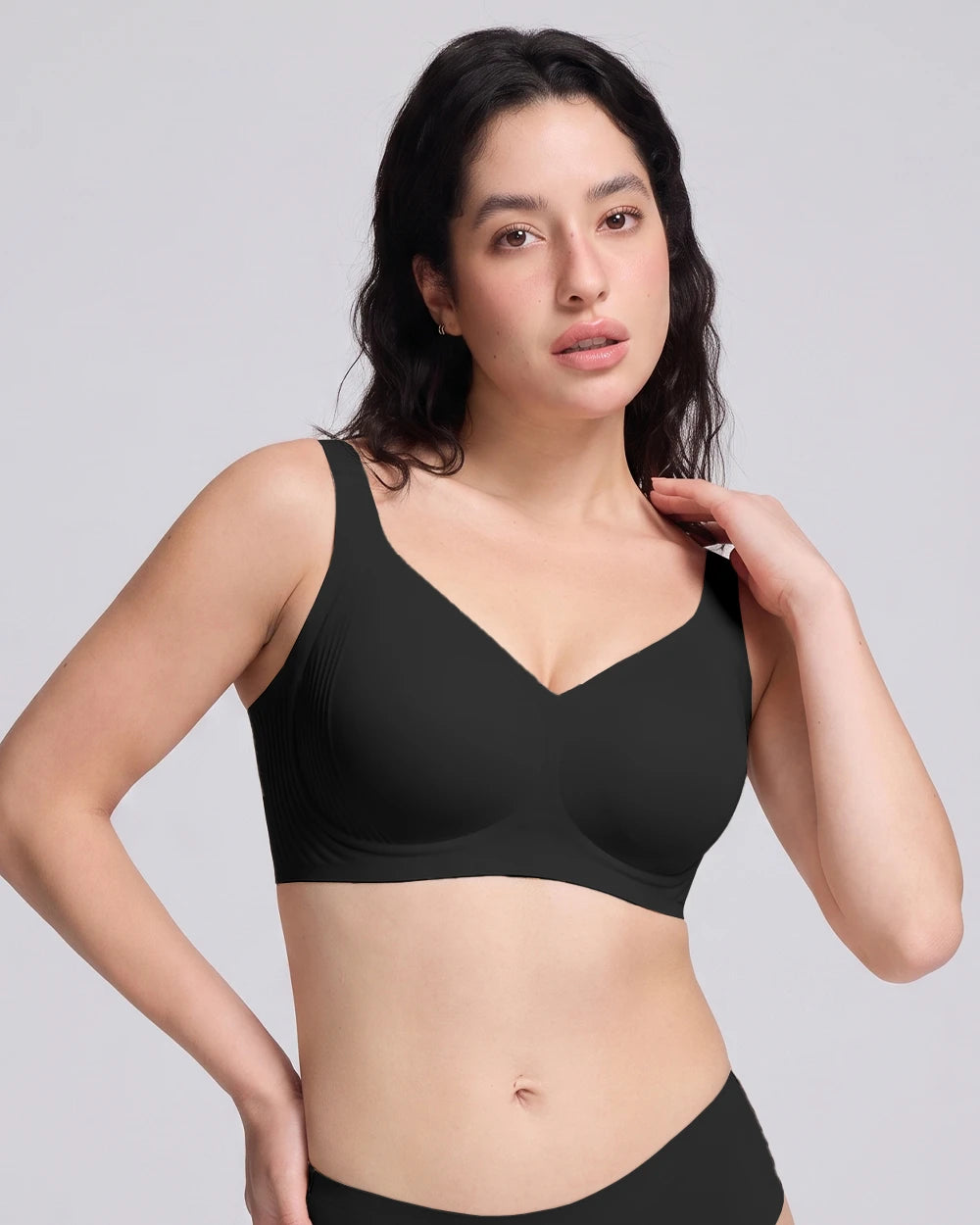A woman modeling a  VRCOMFY black wireless bra with a V-neckline against a solid light gray background.