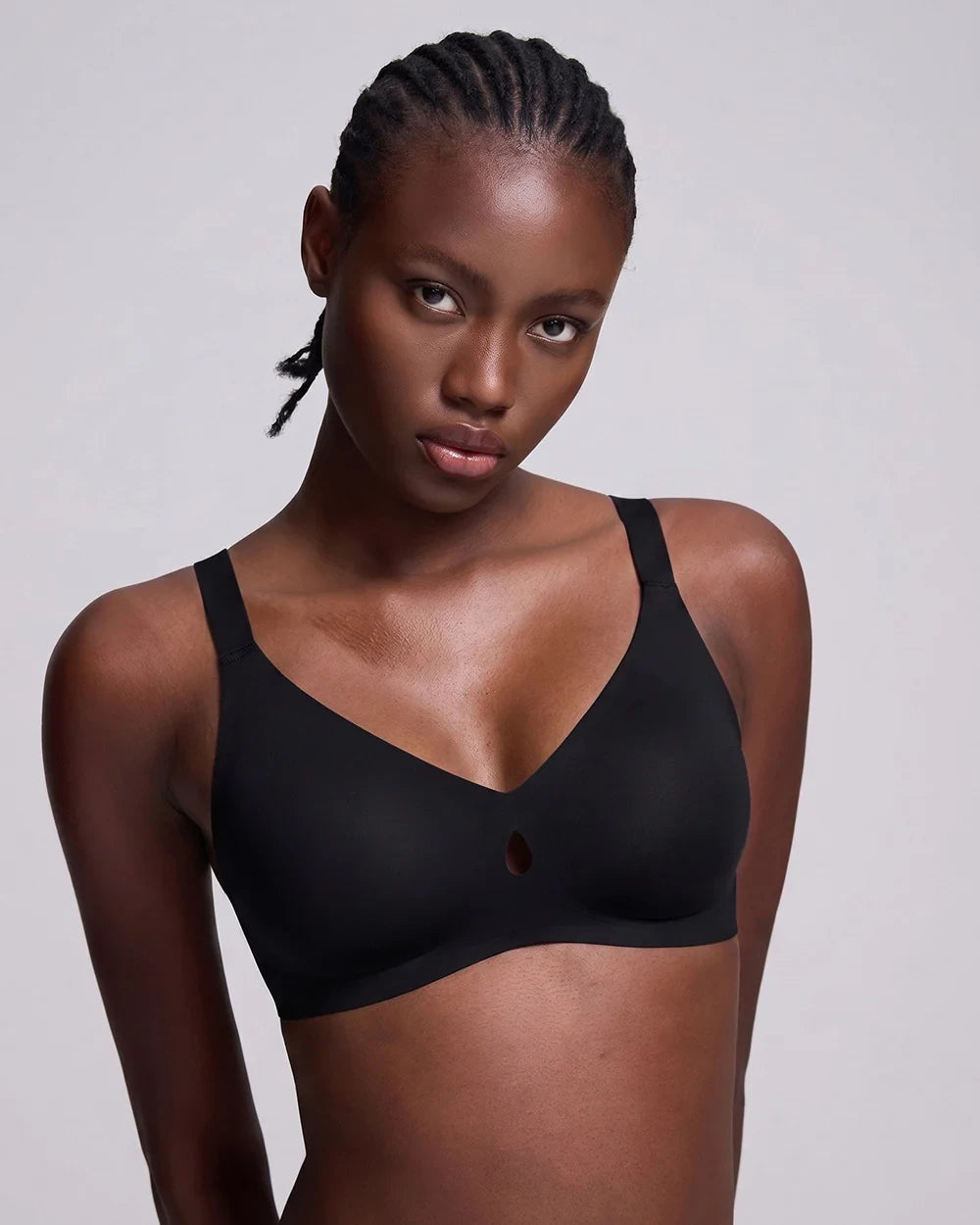 A woman modeling a black wireless bralette with a small cutout at the center, photographed against a neutral light gray studio background.