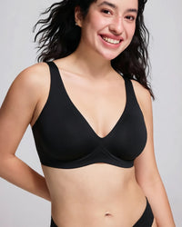 A smiling woman with dark hair wearing a black wireless bra, shown from the chest up against a light gray studio background.