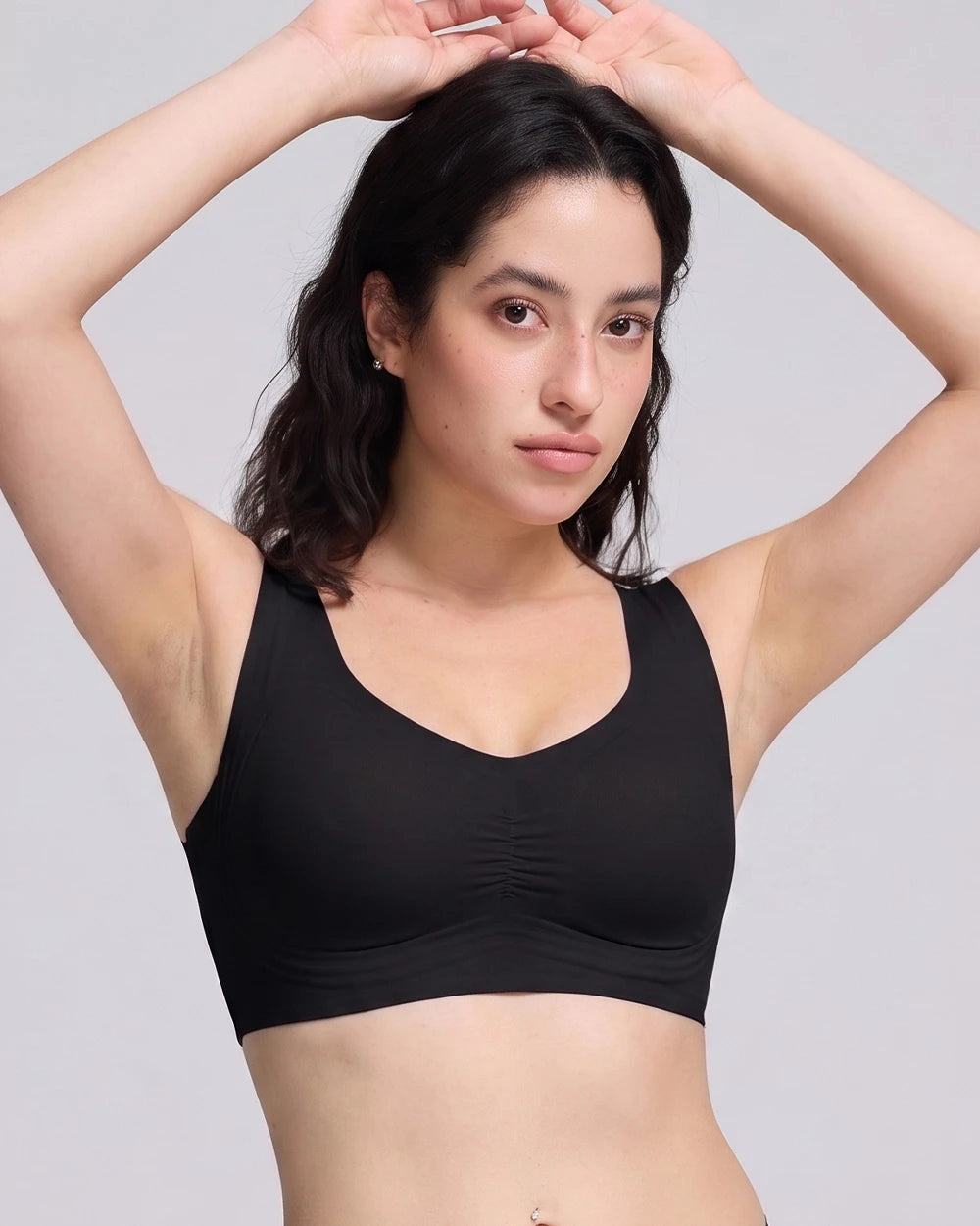 A woman modeling a black wireless sports bra with a gathered underband, posing with her hands in her hair against a light gray background.