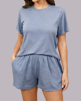 A woman wearing a light blue two-piece loungewear set consisting of a short-sleeve crewneck t-shirt and matching drawstring shorts, shown against a solid gray studio background.