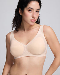 A woman wearing a seamless, light beige/nude wireless bra with molded cups, shown from the chest down against a solid light gray background.