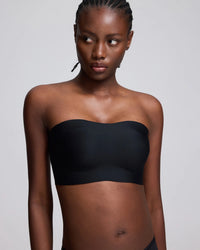 A woman modeling a black strapless bandeau-style bra top against a light gray studio background.