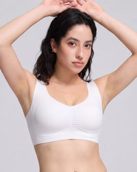 A woman with dark hair modeling a white wireless bralette with a ruched center front, set against a solid light gray background.