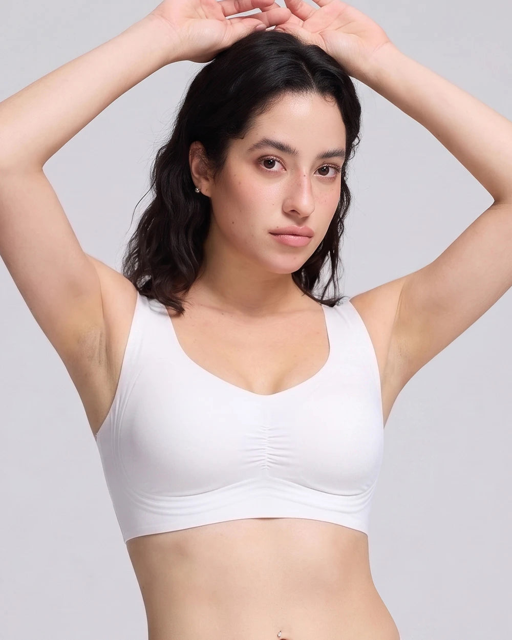 A woman with dark hair modeling a white wireless bralette with a ruched center front, set against a solid light gray background.