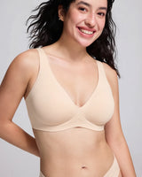 A smiling woman wearing a light beige wireless bra against a plain light gray studio background.