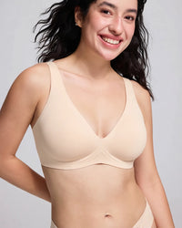 A smiling woman wearing a light beige wireless bra against a plain light gray studio background.