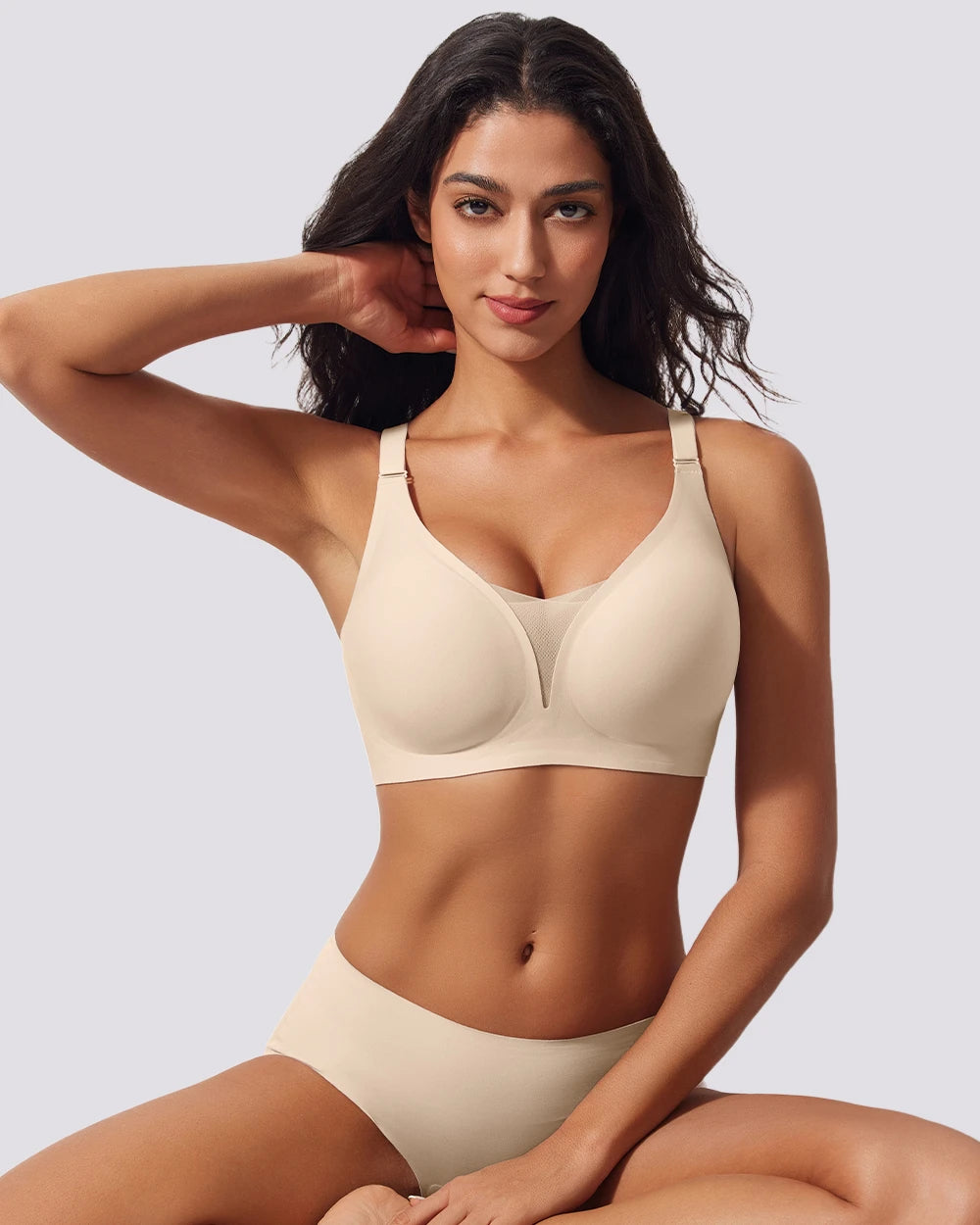 A woman modeling a matching beige wireless bra and high-waisted brief set against a light gray studio background.