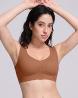 A woman wearing a brown scoop-neck bralette with gathered detailing at the center bust, posing with her hands in her hair against a solid light gray background.